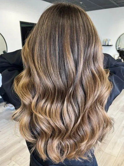 Back view of a woman with long, wavy, brown hair with blonde highlights in a salon setting.