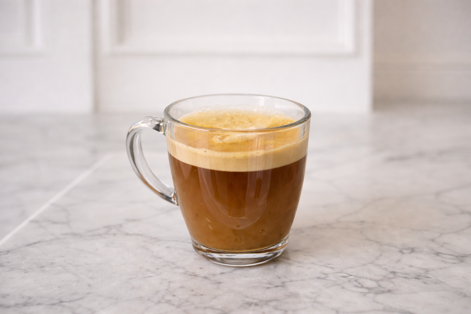A clear glass mug filled with a hot black coffee topped with foam, placed on a white marble countertop.