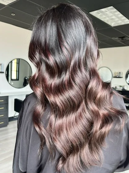 Woman with long, wavy hair with a dark top and a salon background featuring mirrors and chairs.
