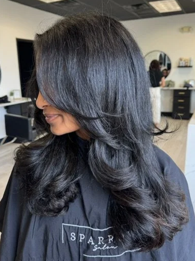 A woman with long, black, wavy hair sitting in a salon chair, with salon equipment visible in the background.