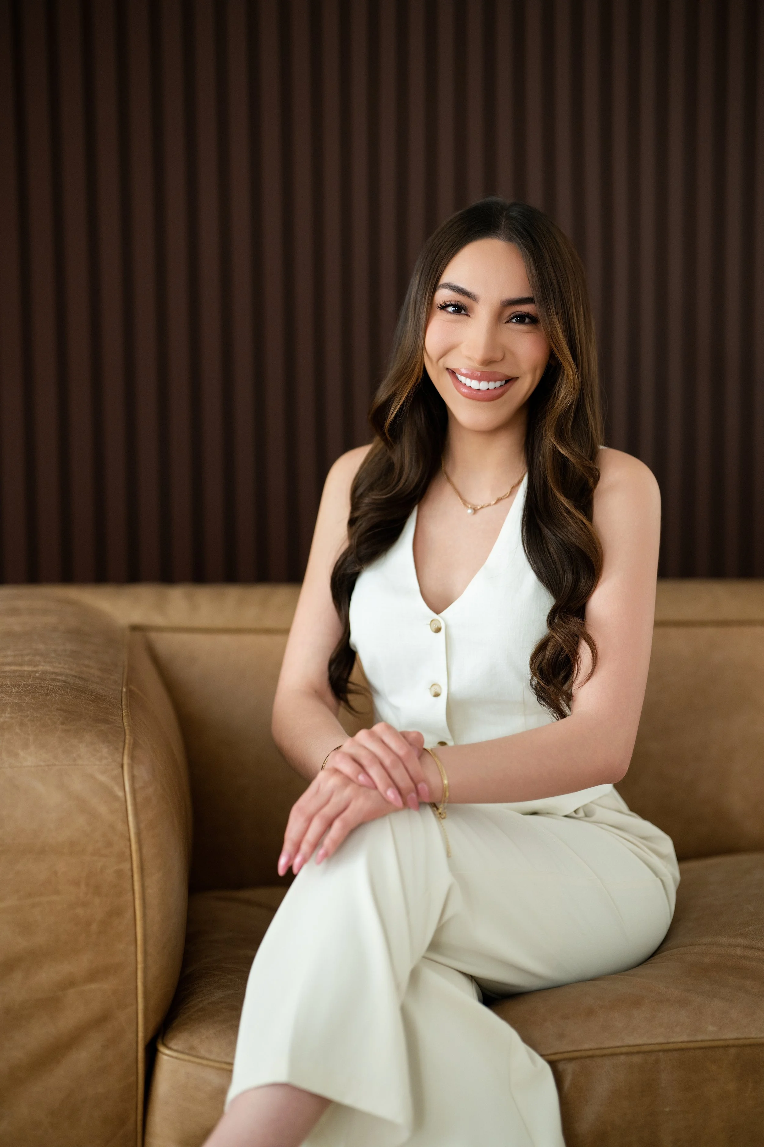 A young woman with long wavy dark brown hair, wearing a white sleeveless button-down dress, is sitting on a brown leather couch. She is smiling, showing her teeth, and holding her hands together on her lap. She is accessorized with a delicate gold necklace, a bracelet, and a ring, with a background featuring dark wooden paneling.