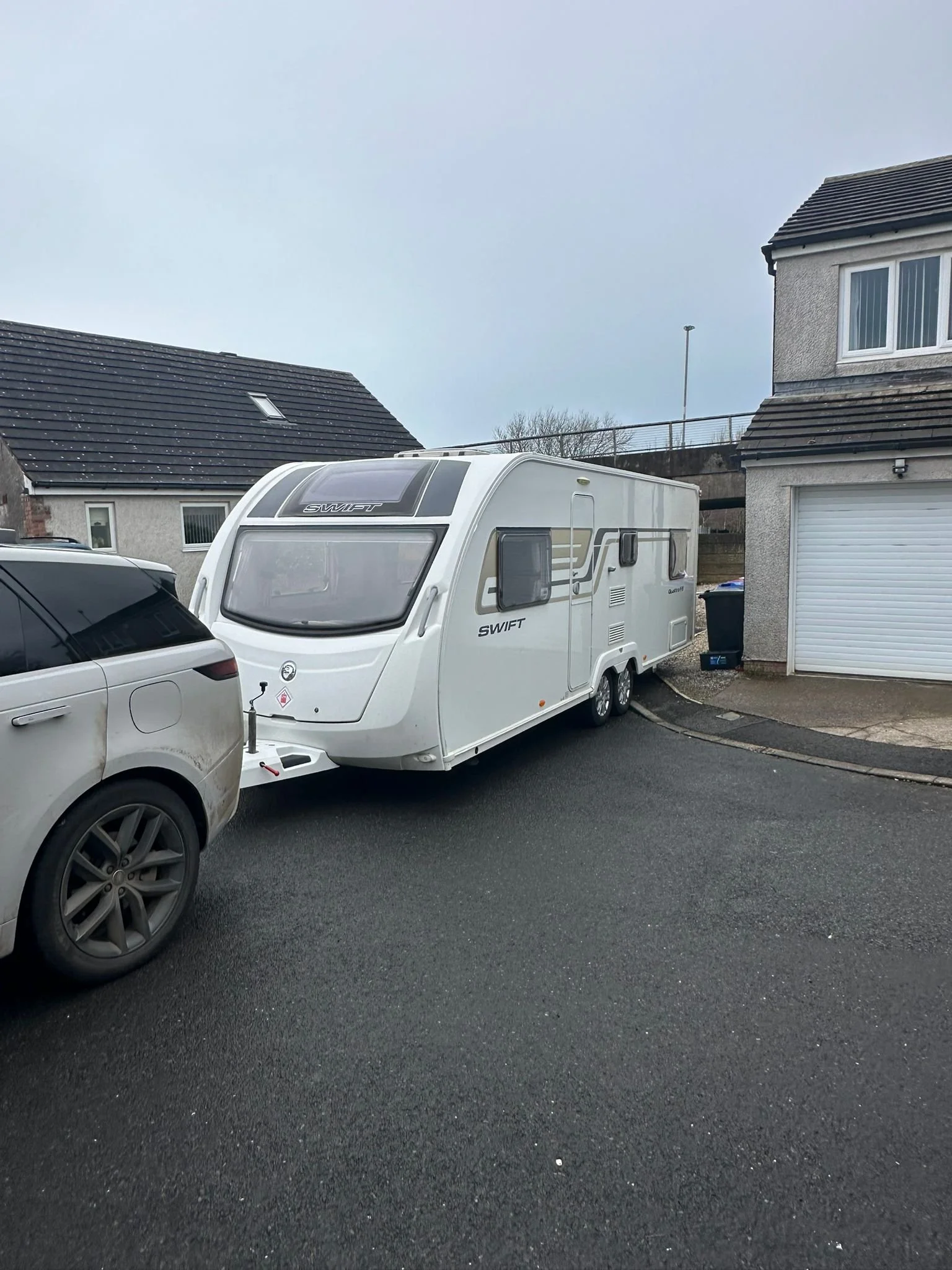 A white caravan attached to a gray car parked on a residential street with houses and a garage in the background.