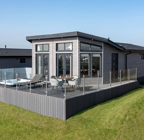 Modern house with a glass-enclosed balcony, outdoor furniture, and hot tub overlooking a grassy area.