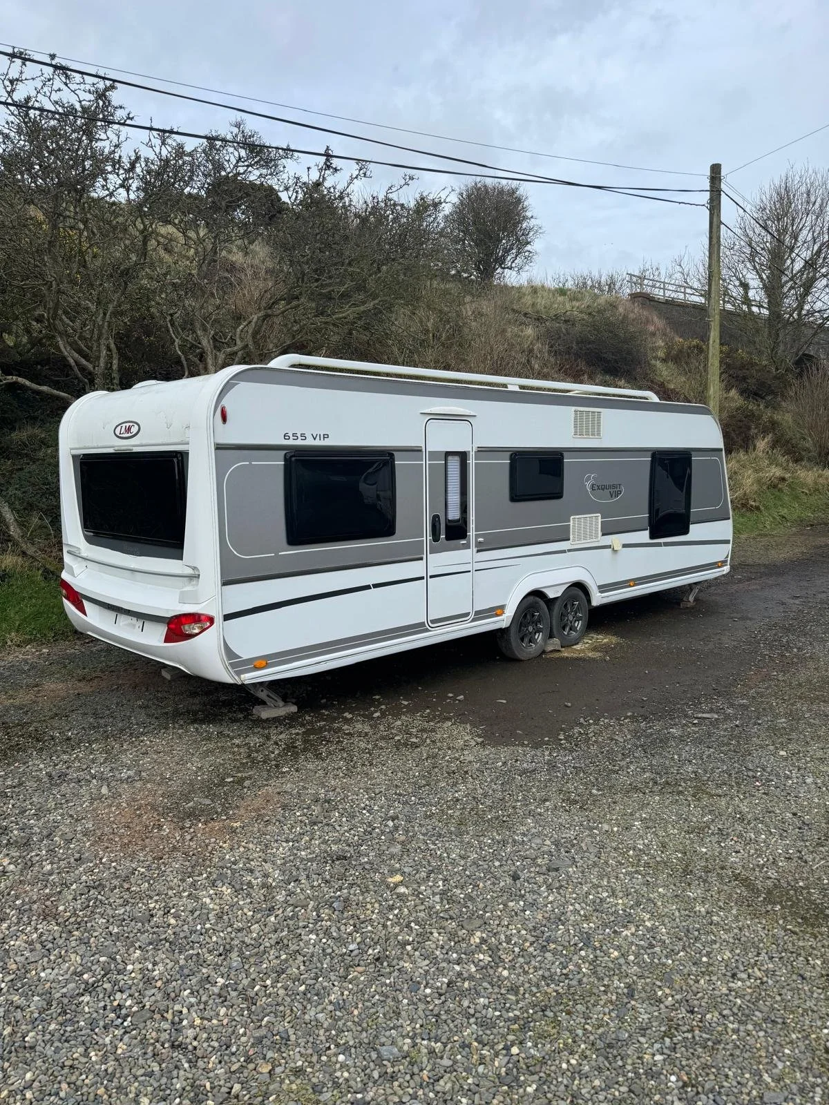 A white travel trailer parked on a gravel lot with a hillside and trees in the background.