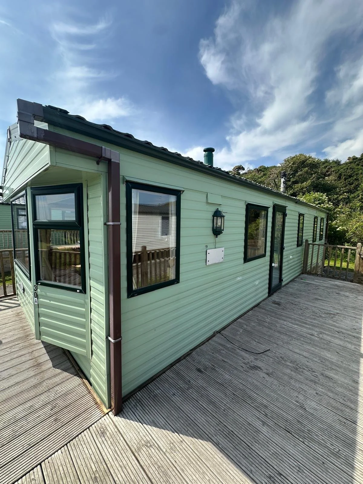 A light green tiny house on a wooden deck under a blue sky with clouds, with surrounding trees and mulch.