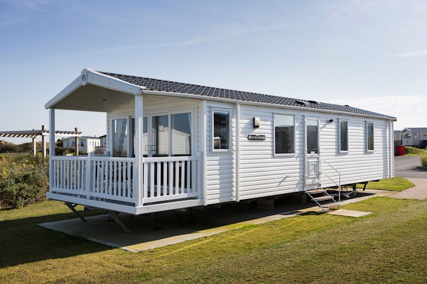 White mobile home with a porch, situated on a grassy area with a concrete foundation, in a neighborhood with other similar homes visible in the background.