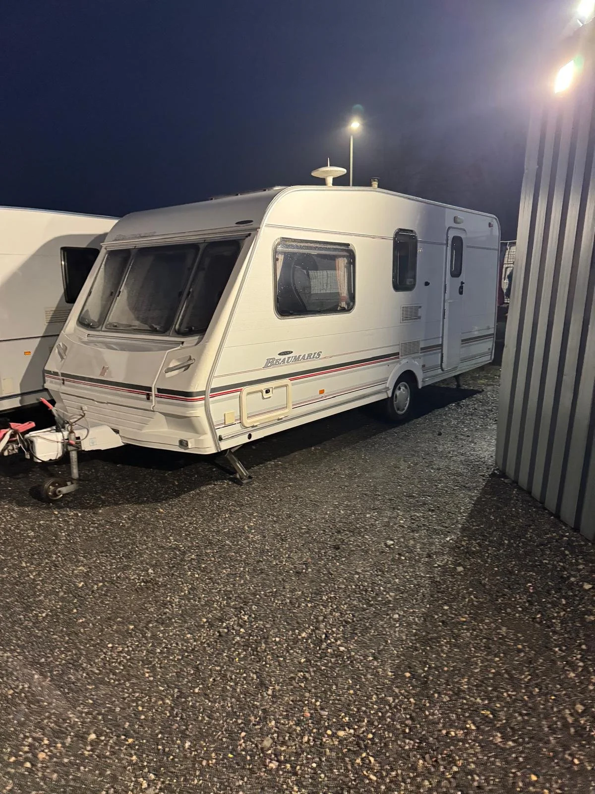 A white travel trailer parked on a gravel lot at night, illuminated by nearby lights.