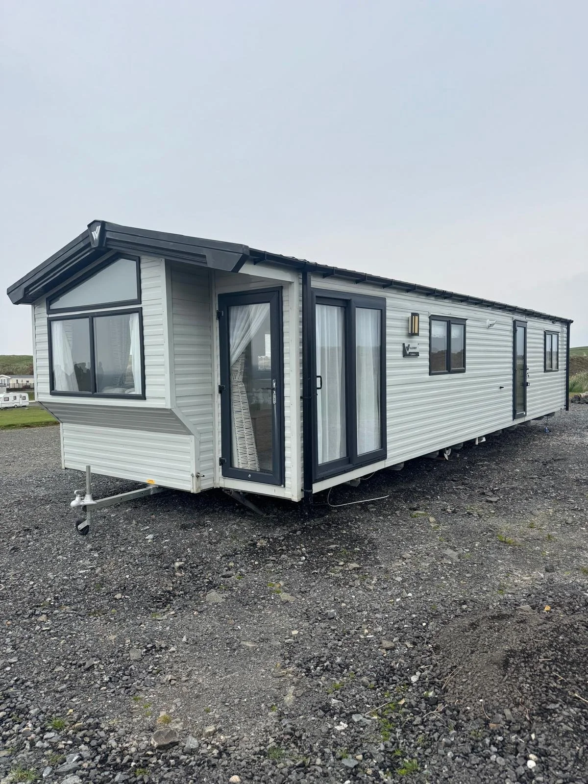 A white mobile home with black trim on a gravel lot under an overcast sky.