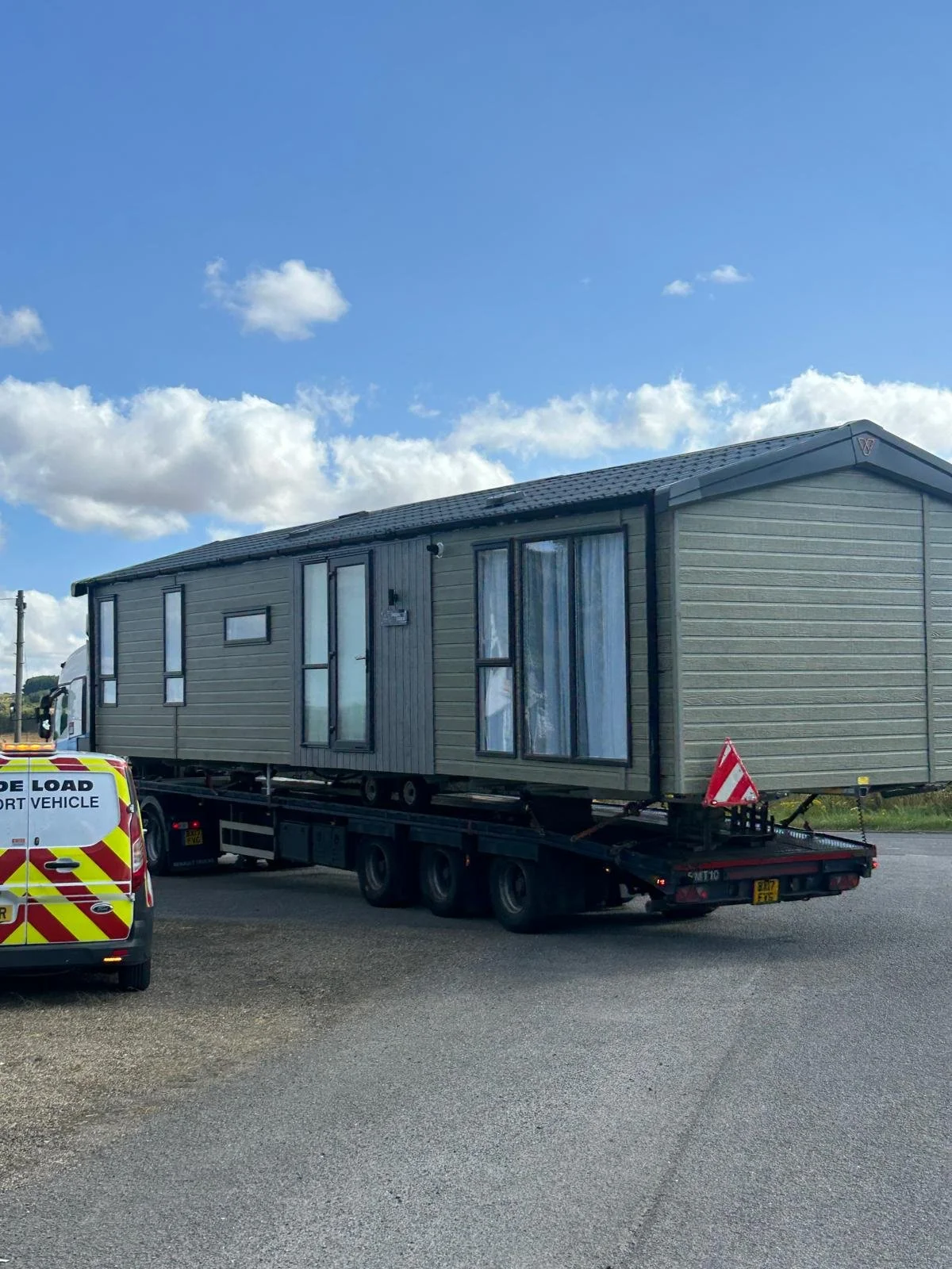 A large mobile home being transported on a flatbed truck on a road, with a police or emergency vehicle nearby, under a partly cloudy sky.