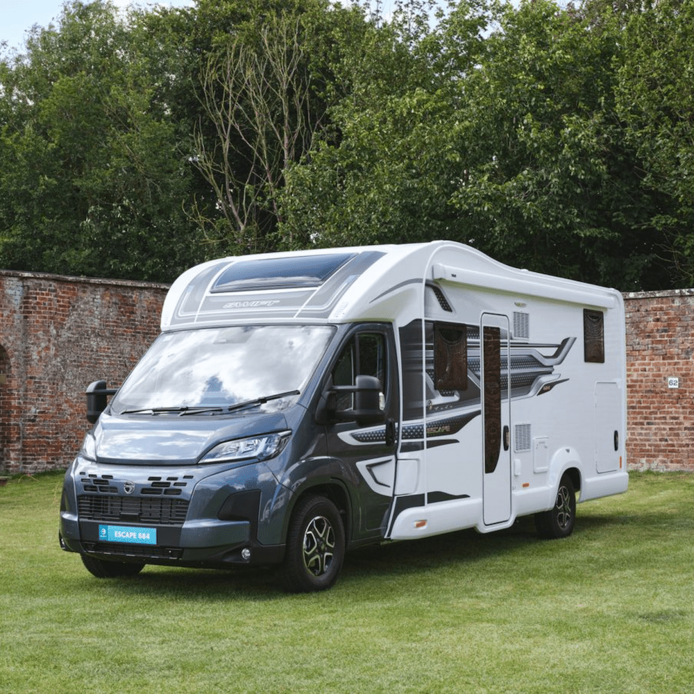 A modern white and gray camper van parked on a grassy area with a brick wall and green trees in the background.