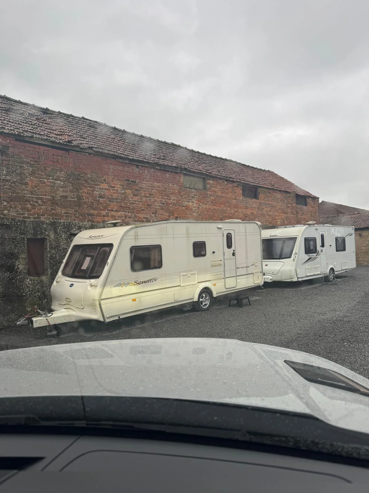 Two RV trailers parked on a gravel lot in front of a brick building with a sloped roof, seen through a vehicle's windshield.