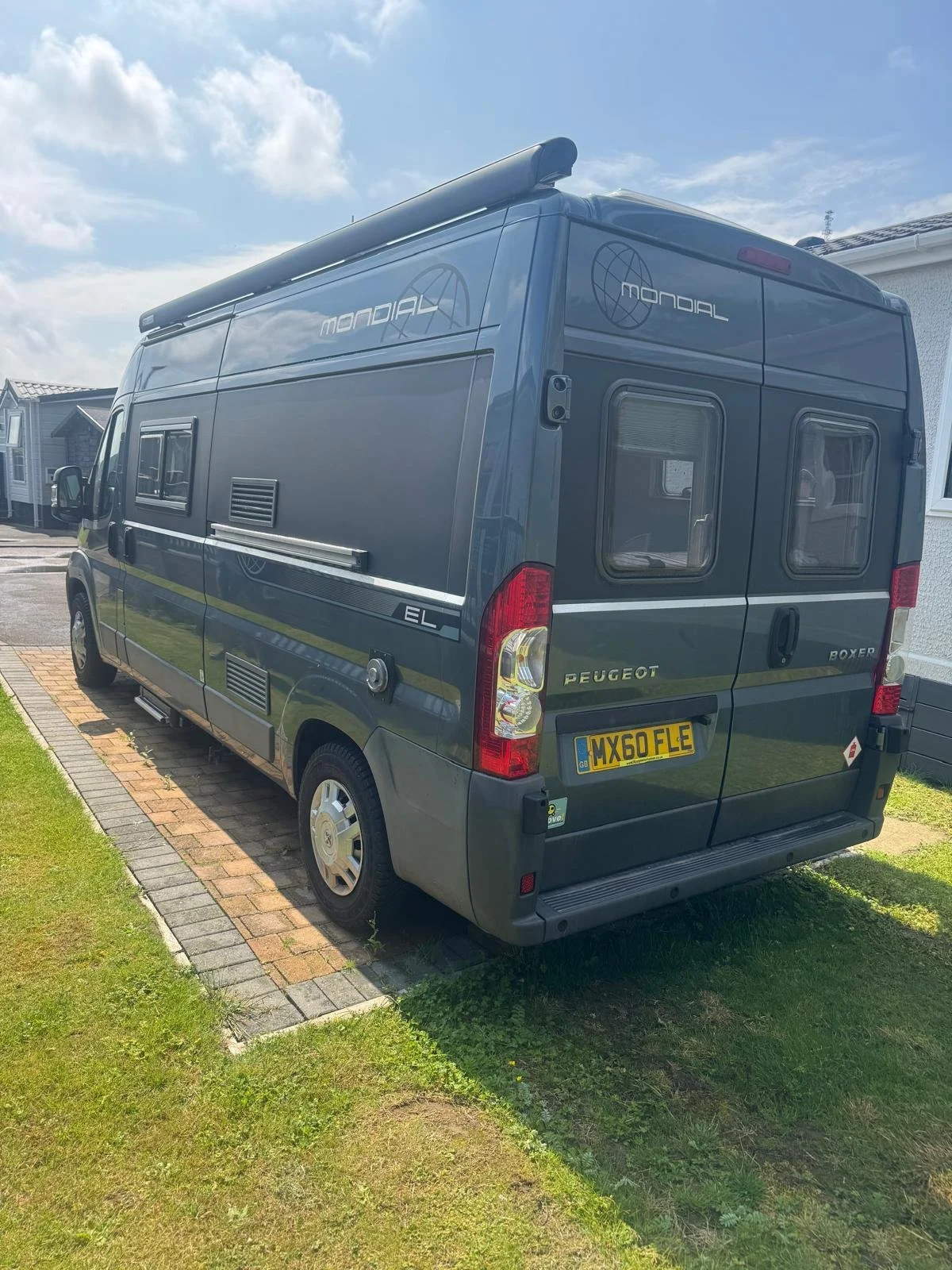 Gray Peugeot Boxer camper van parked on a driveway next to a house with a grassy lawn and brick paving.