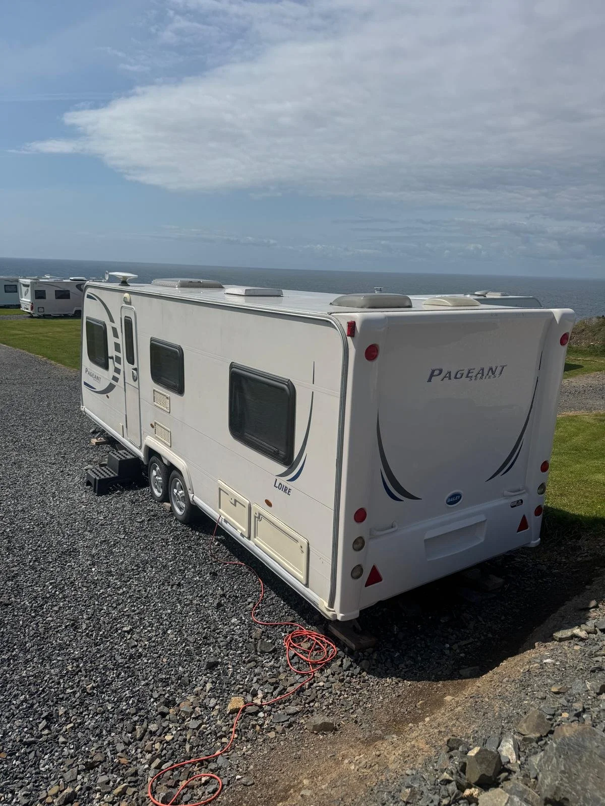 A white recreational vehicle (RV) parked on gravel with the ocean in the background and cloudy sky overhead.