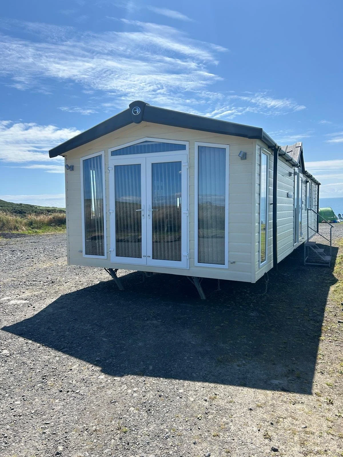 A beige and black mobile home with large glass doors and windows, set on a gravel surface outdoors under a blue sky with clouds.