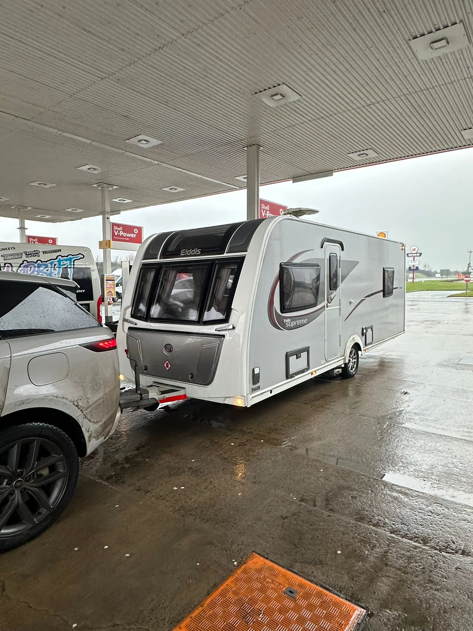 A white travel trailer hitched to a gray SUV parked under a gas station canopy on a rainy day.