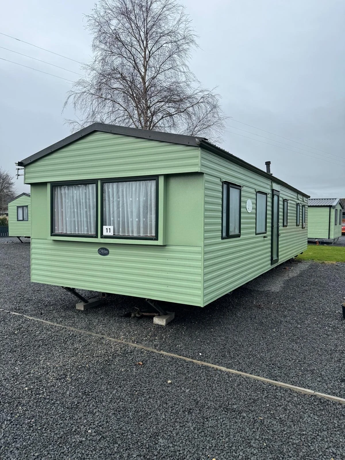 A green mobile home with black-trimmed windows and a small white number '11' sign in the front window, set on a gravel lot with other similar mobile homes, overcast sky, and a leafless tree in the background.