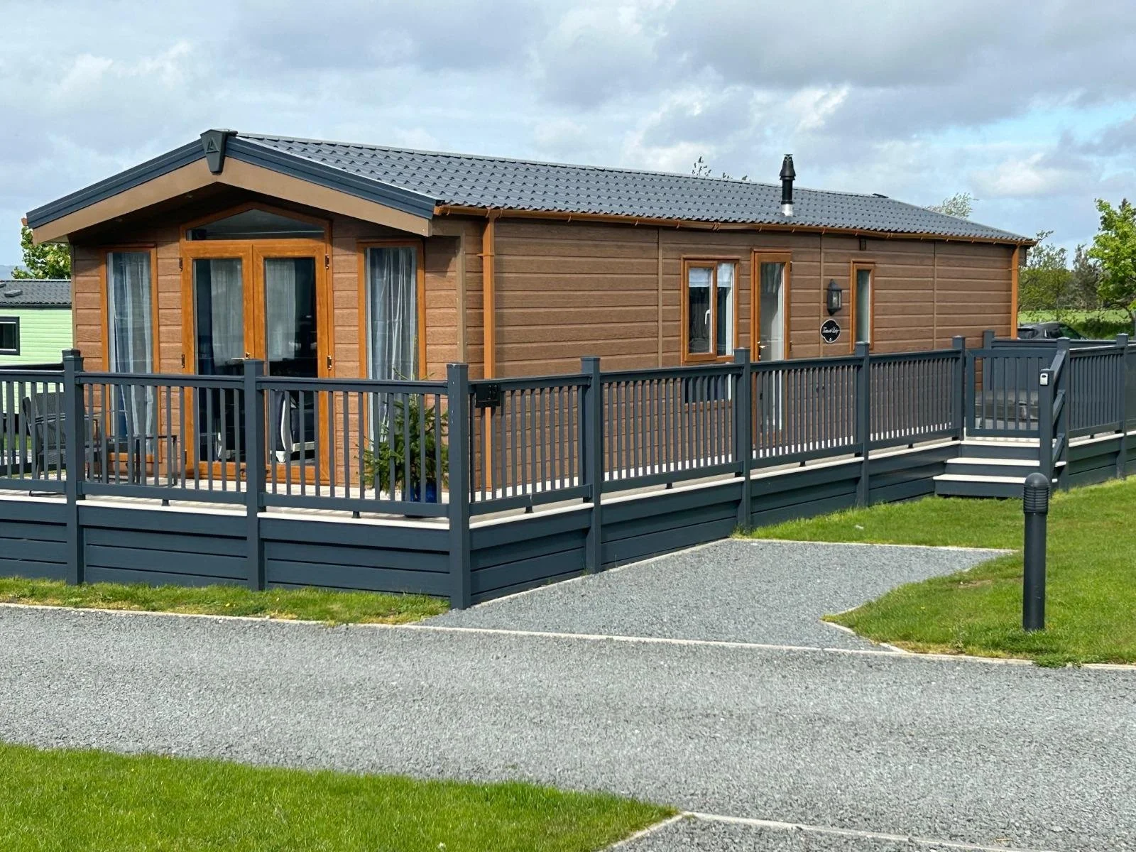 A wooden house with a gray roof, large glass doors, and windows, surrounded by a dark gray fence with a ramp, green grass, and a gravel pathway.