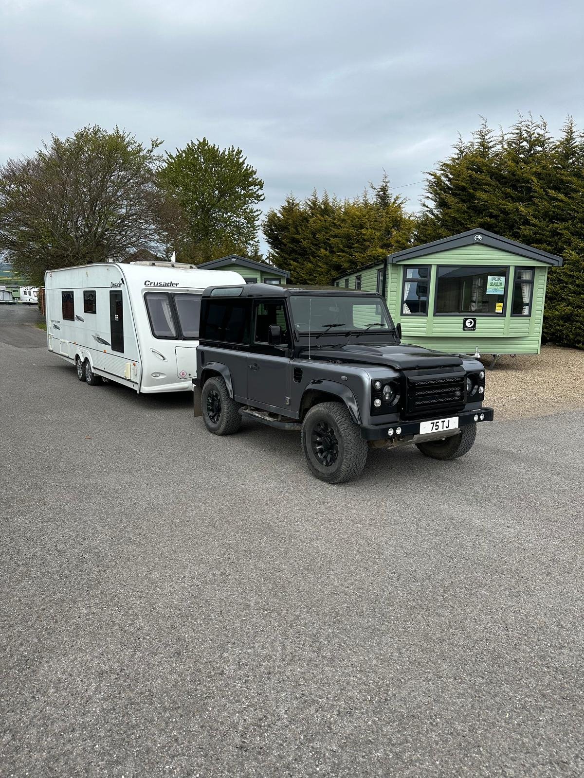 A black Land Rover Defender parked behind a white caravan on a gravel lot with green mobile homes and trees in the background.
