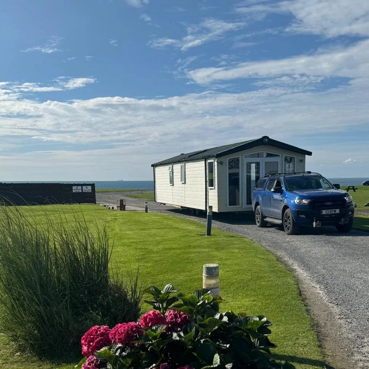 A small white house on a gravel path with a blue car parked beside it, green grass, pink flowers, and a view of the ocean under a partly cloudy sky.