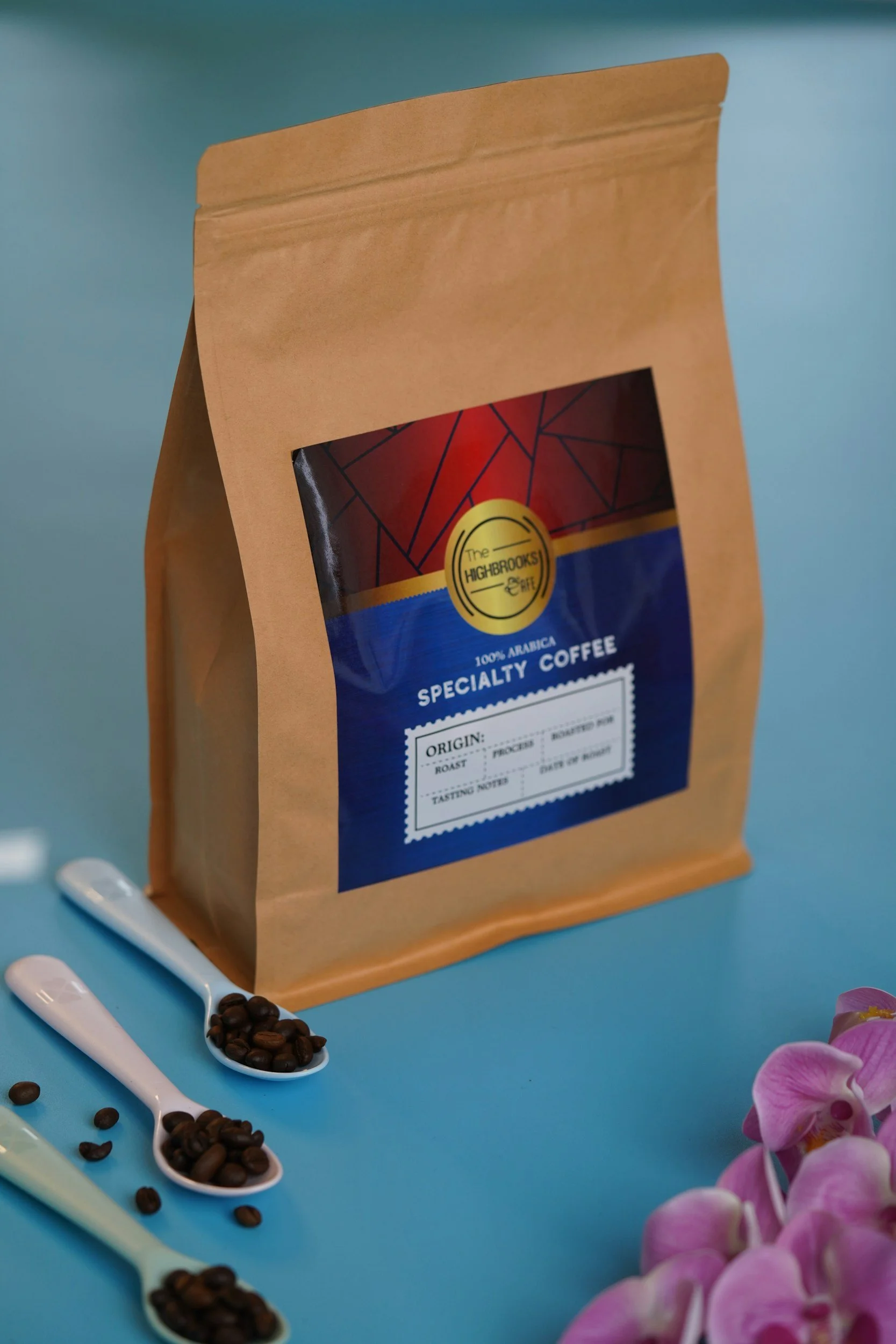 Paquete de café de origen árabe, marca The High-Brooks, en una bolsa de papel kraft con etiqueta azul y roja, en un fondo azul. Rodeado de cucharas con granos de café y flores rosadas.