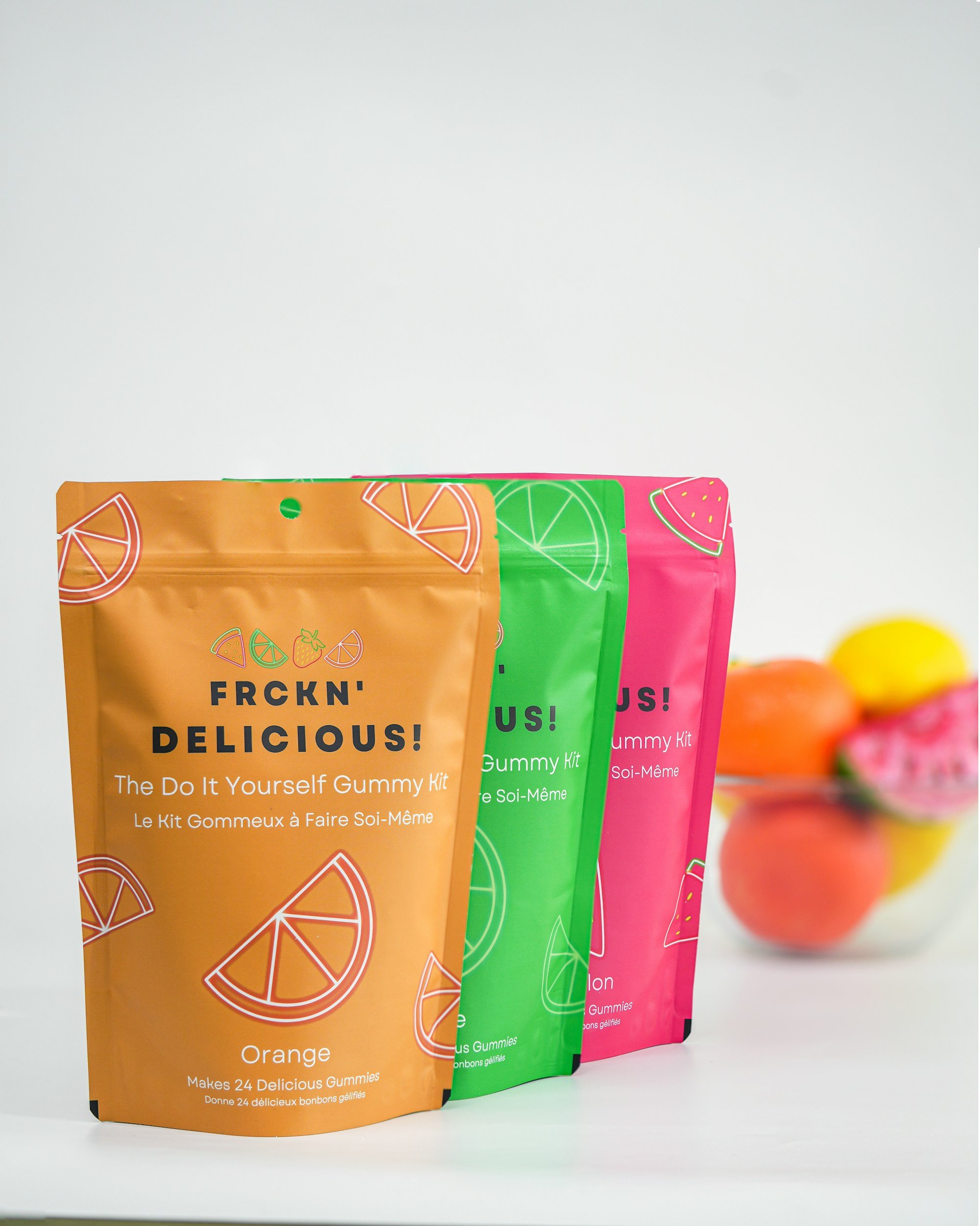 Paquetes de gomitas en colores naranja, verde y rosa con diseños de frutas en la etiqueta, en primer plano, con una jarra con frutas en el fondo.