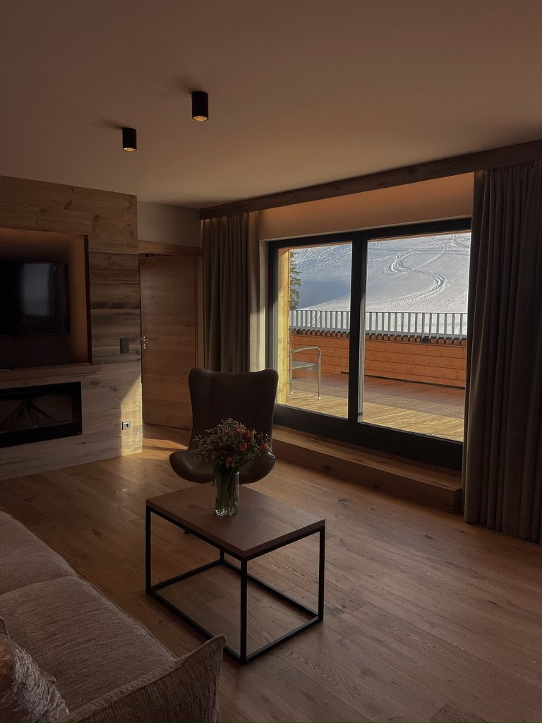 Living room with wooden flooring, a glass door leading to a balcony with snow-covered slopes, a TV in a wooden wall niche, a black armchair with a floral cushion, a coffee table with a vase of flowers, and curtains framing the view.