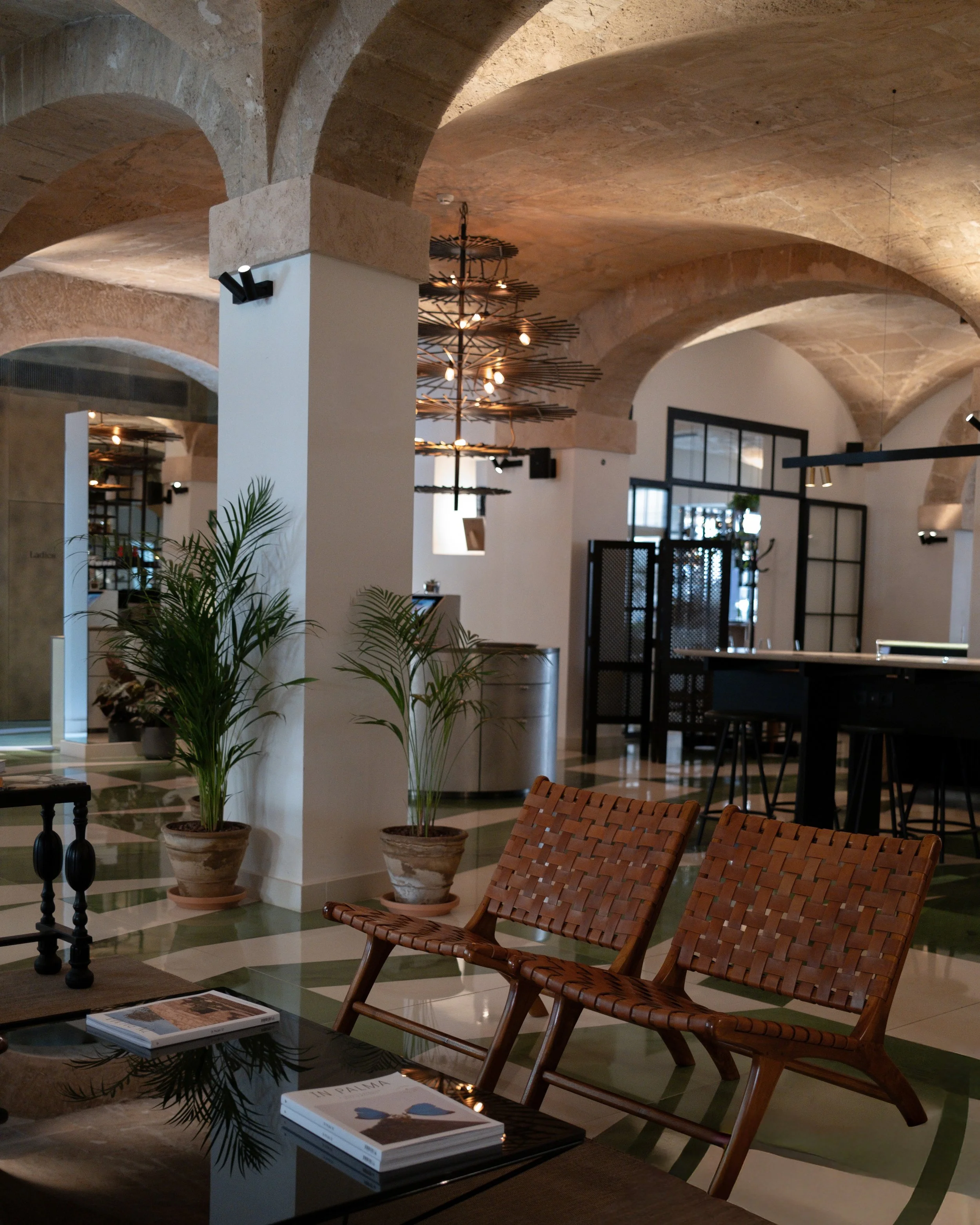 Interior of a modern, stylish lounge or cafe with arched stone ceilings, potted plants, woven chairs, a black coffee table with magazines, and a distinctive chandelier hanging from the ceiling.