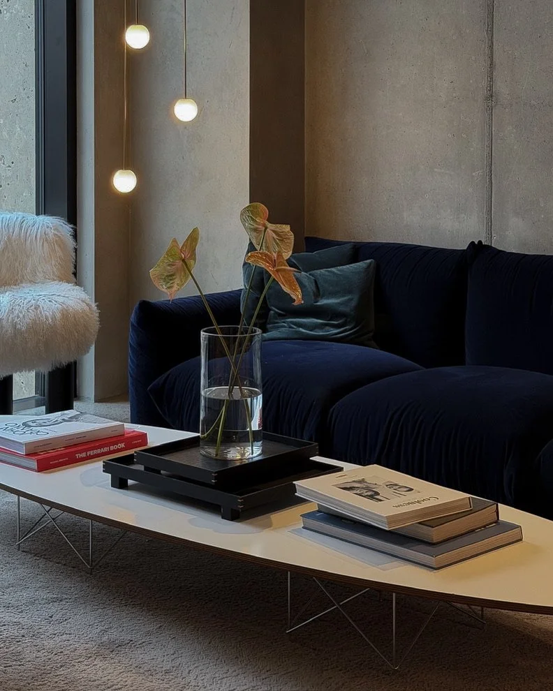 Modern living room with a navy blue sofa, a white coffee table with magazines and a glass vase with leaves, a textured cream and fluffy chair, and hanging globe pendant lights near a large window with a concrete wall.