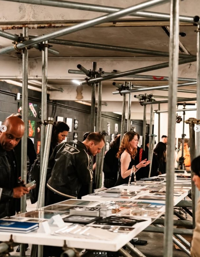 People shopping at an art gallery, browsing artworks and photographs on display tables amidst scaffolding.