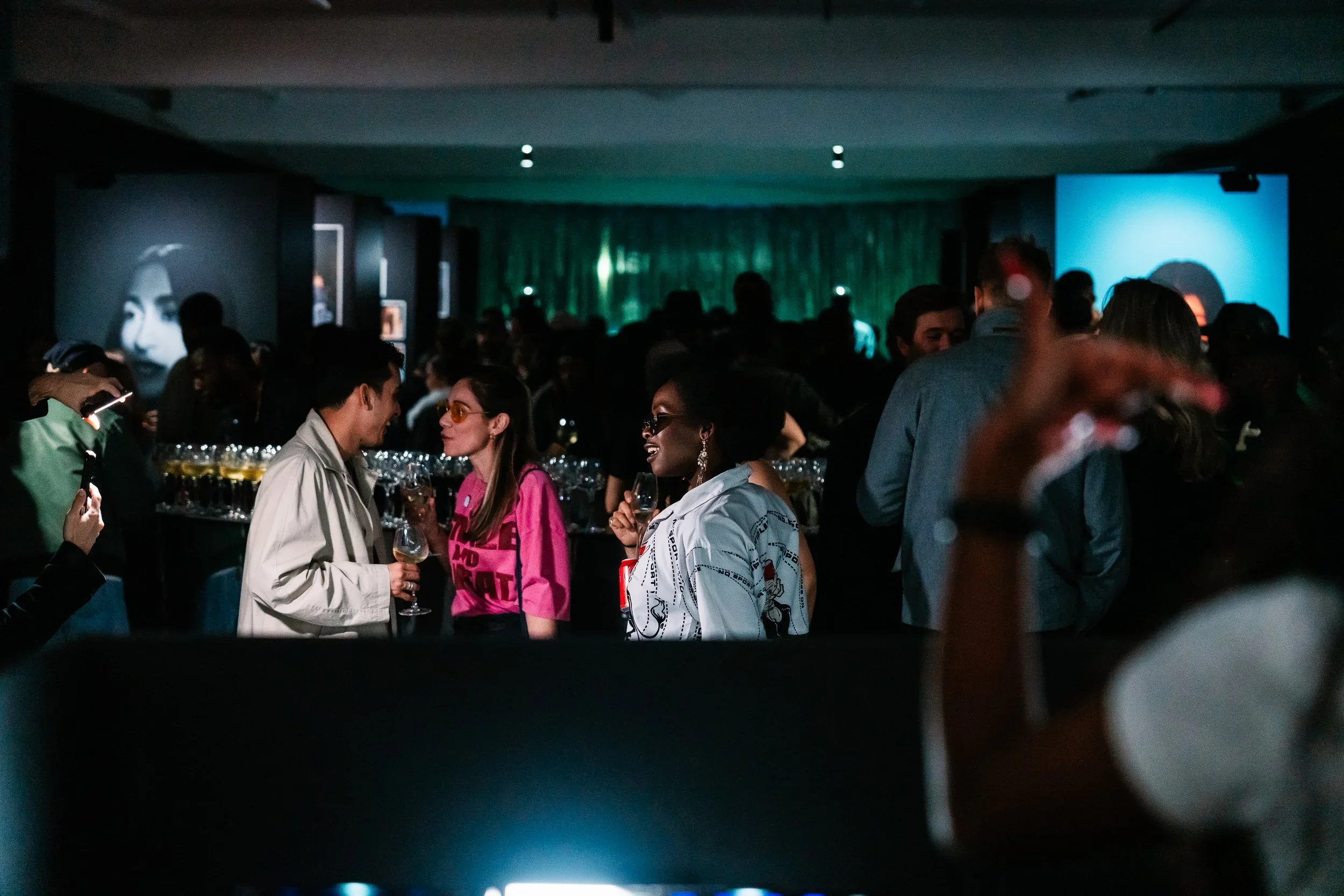 People socializing at a dimly lit event or party, with some holding drinks, in a room with dark walls and projection screens showing faces and images.