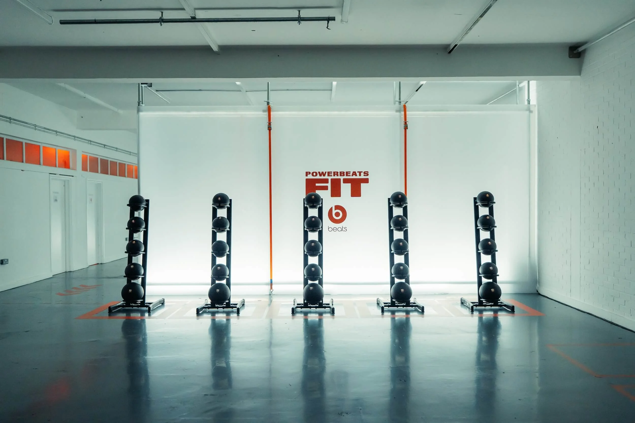 Empty fitness studio with six vertical racks holding exercise balls in front of a wall with red Powerbeats FIT and Beats logos