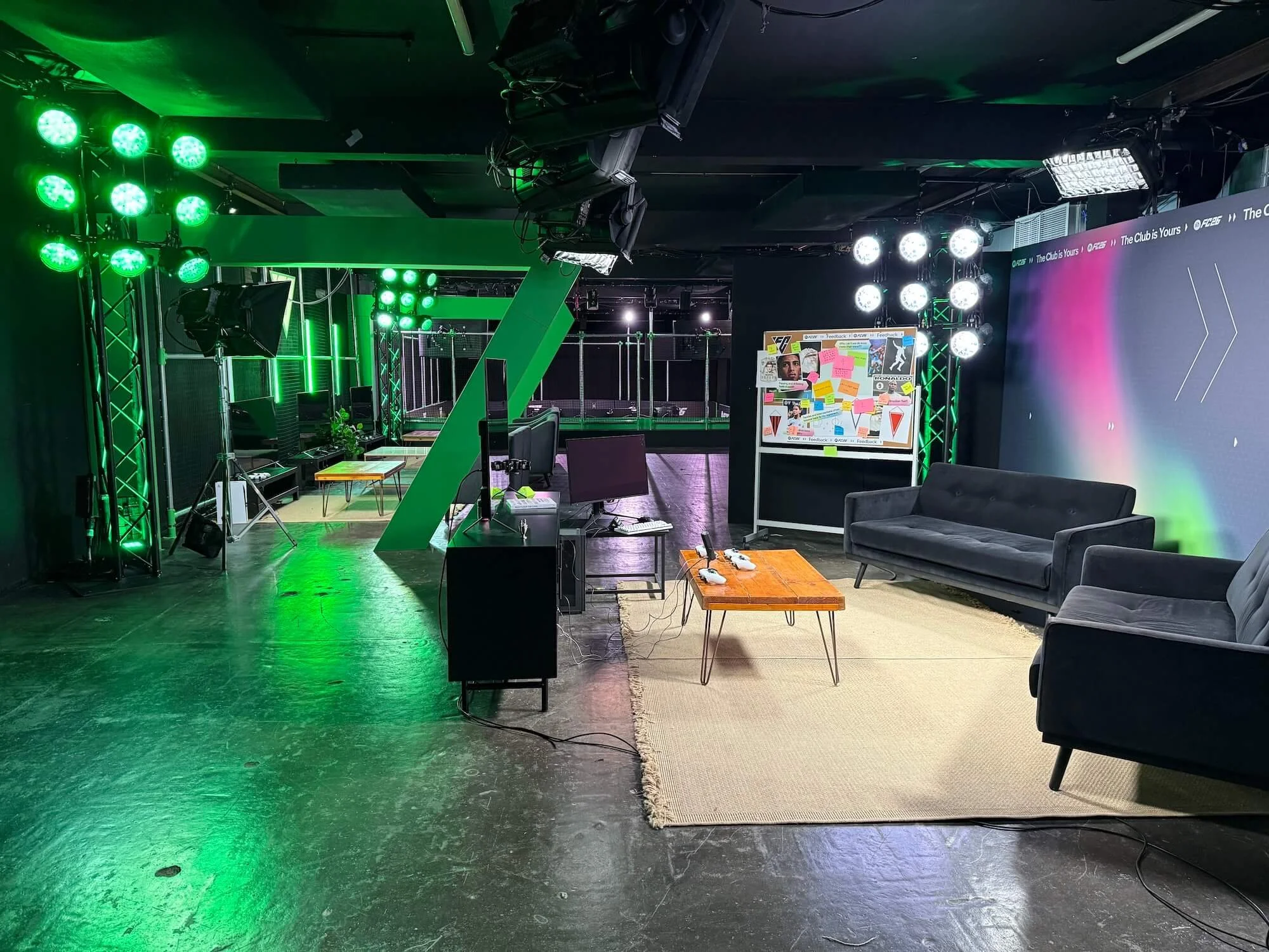 Interior of a modern event or projection space with black flooring and seating areas, illuminated stage lighting in green and white, a large screen, desks with computers, and a whiteboard with sticky notes.