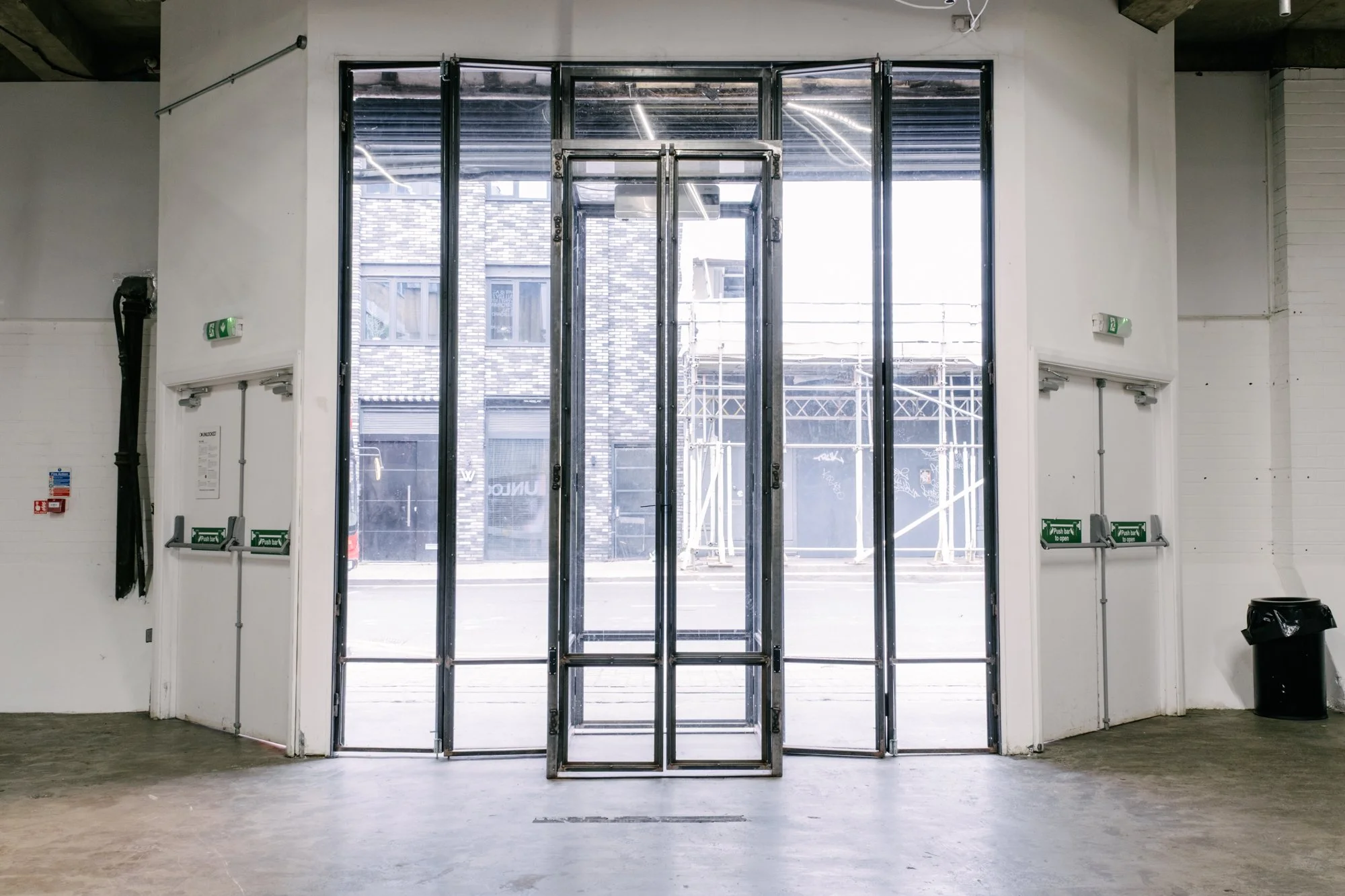 Empty interior with glass double doors leading outside, visible buildings and street through the doors, white walls, fire exit signs, black trash can, and some black pipe on the left wall.
