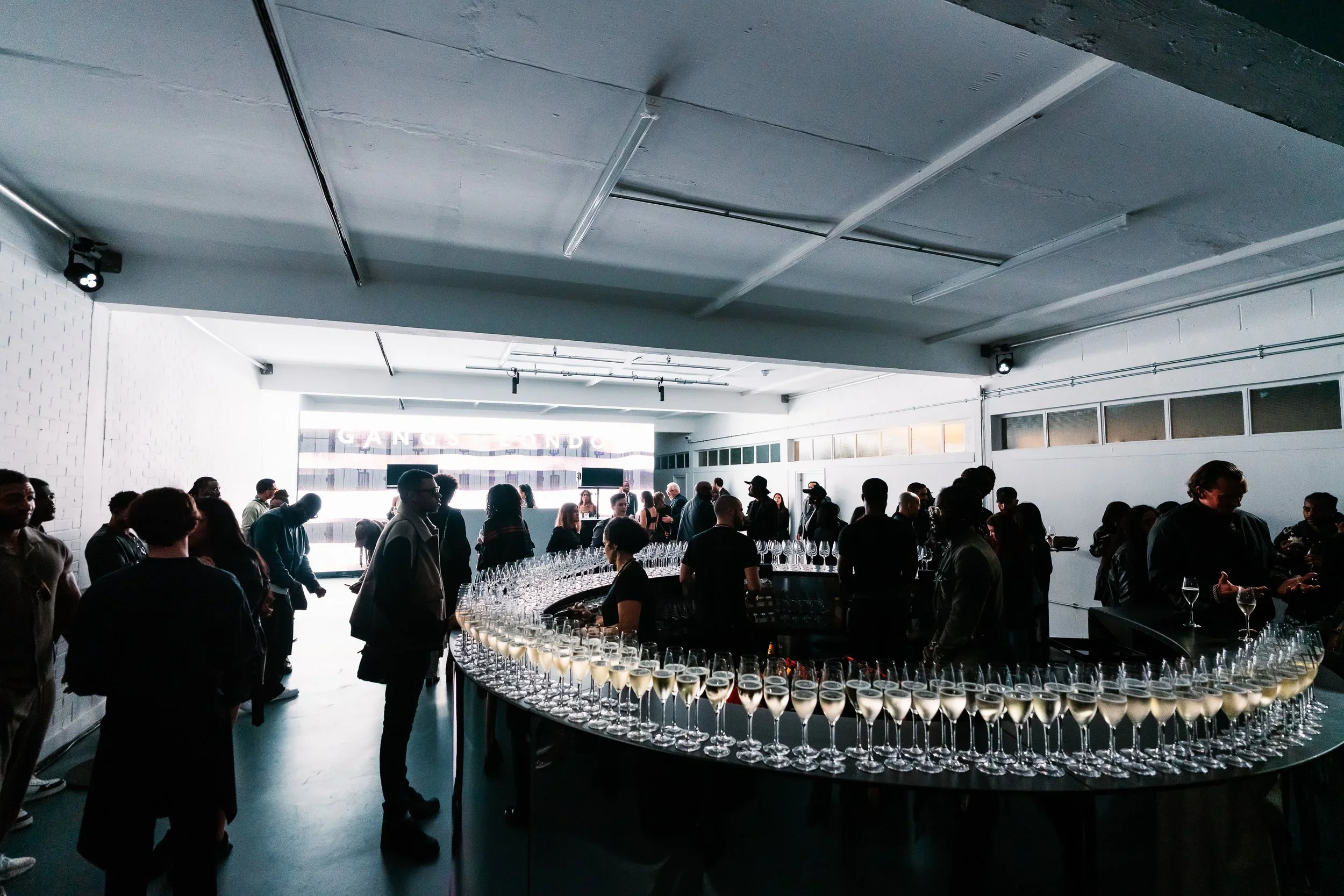 A crowded indoor event with people mingling around a circular table filled with glasses of champagne or sparkling wine, with some people holding drinks and others chatting, in a room with white walls and large windows letting in light.