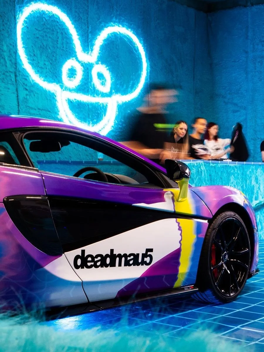A sports car with a vibrant paint job featuring purple, black, white, yellow, and blue colors, parked inside with a neon mouse head sign on the wall behind it. The car has the text "deadmau5" on the side. There are several people in the background, and the floor is covered with blue tiles.
