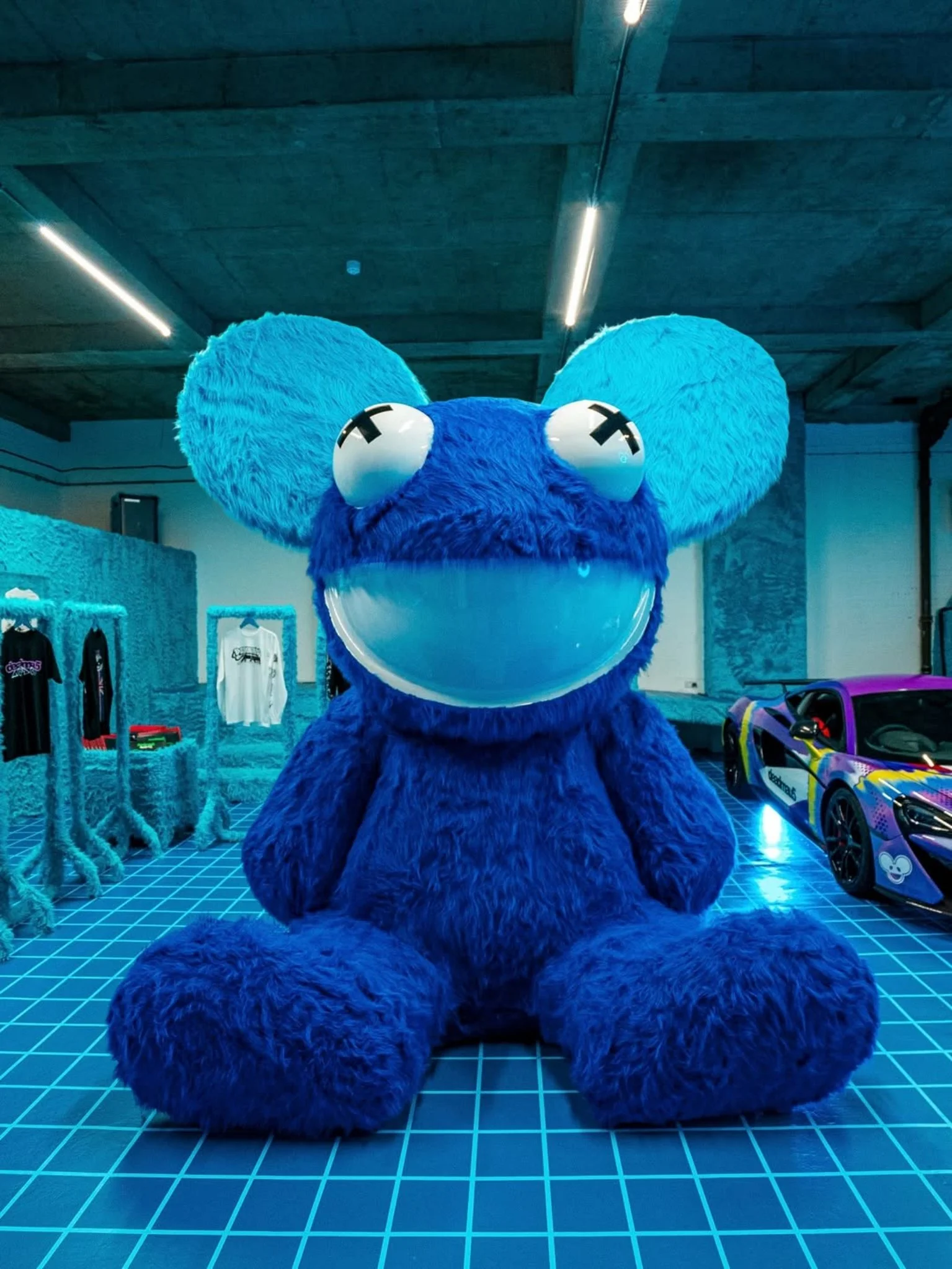 A large, blue, furry mouse print statue with oversized ears and googly eyes, sitting on a blue grid floor in an indoor space with cars and clothing displays.