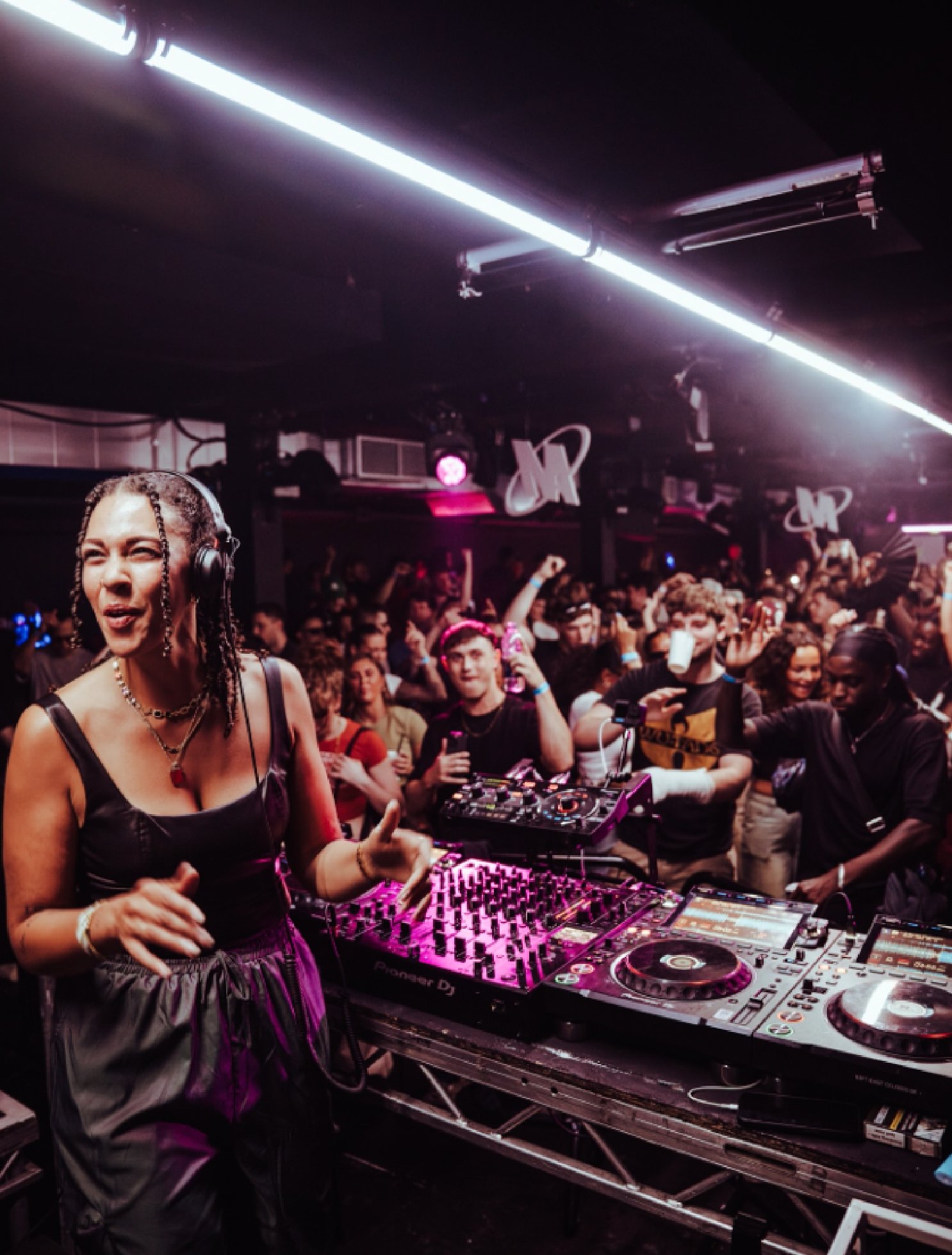 A female DJ performing at a crowded nightclub with colorful lighting, surrounded by dancing guests.