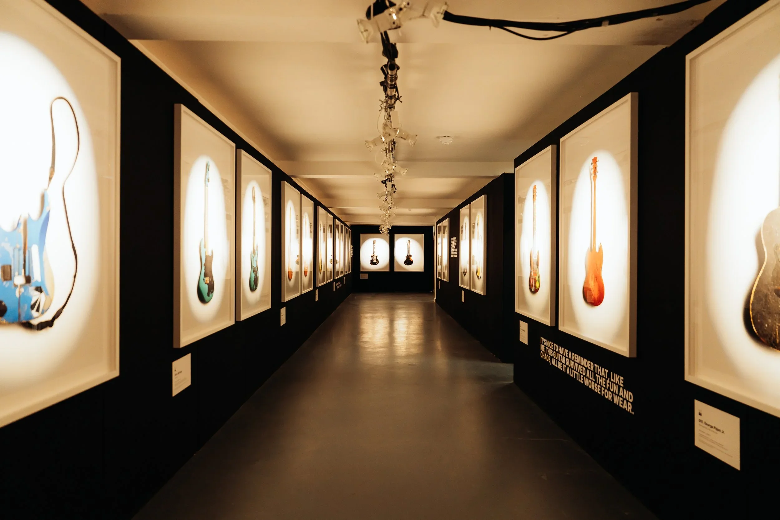 An art gallery with black walls and white-framed pictures of electric guitars on the walls. The gallery has a central track lighting system on the ceiling, illuminating the artwork.
