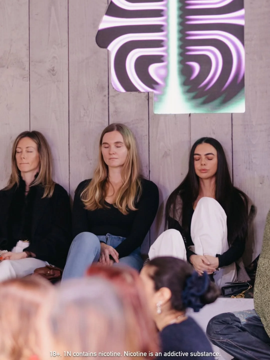 Three women sitting side by side against a wooden wall at a meditation or mindfulness event, with their eyes closed, in front of an abstract purple, white, and black neon sign.
