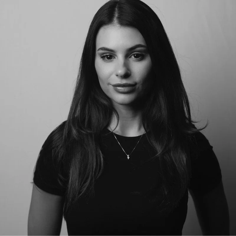 Black and white portrait of a young woman with long dark hair, wearing a black top and a delicate necklace, looking directly at the camera.