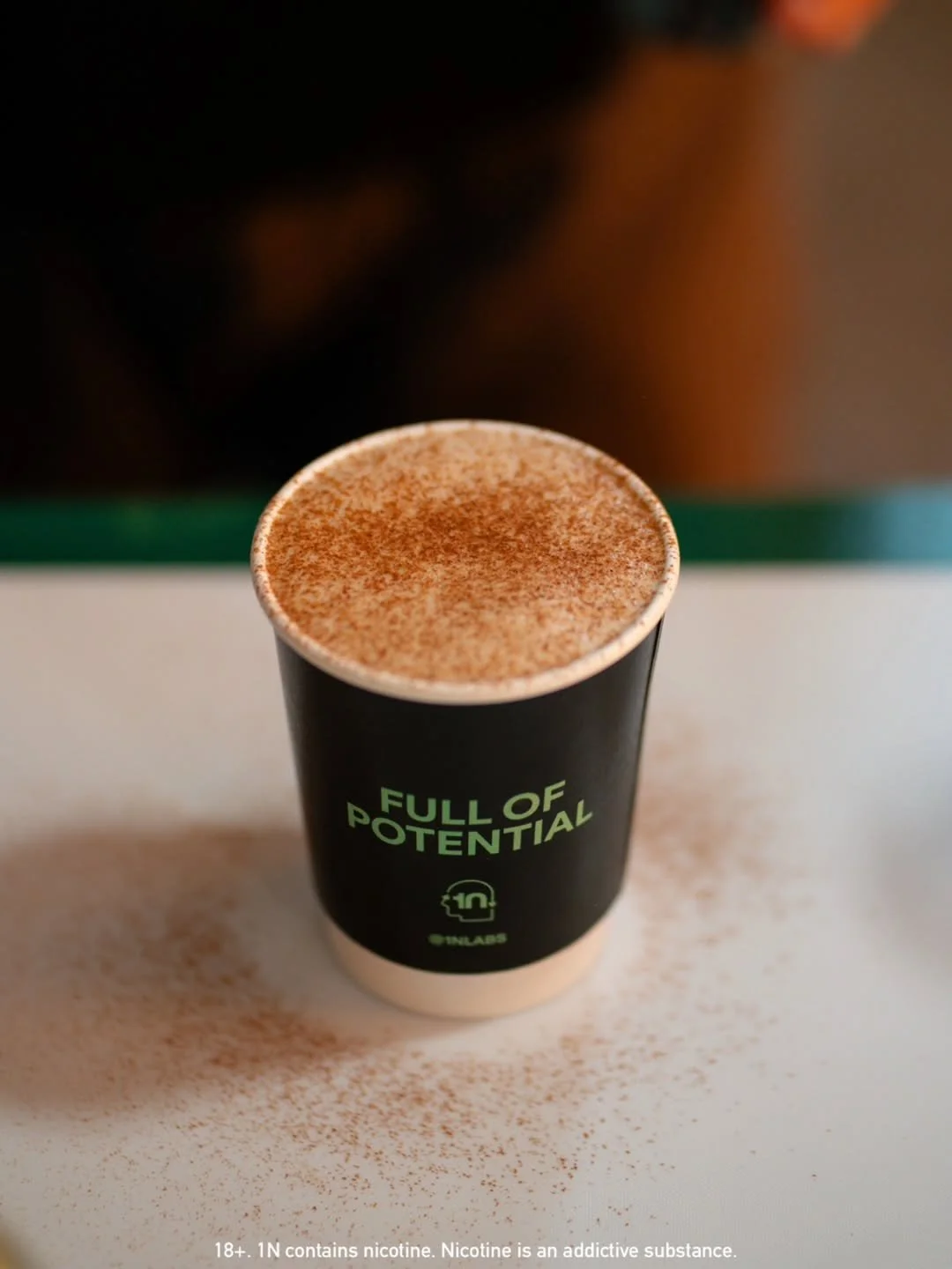 A black paper coffee cup with green text that says 'FULL OF POTENTIAL' and the number 10 in a circle, filled with a frothy beverage topped with cocoa powder, resting on a white surface with cocoa powder sprinkled around.
