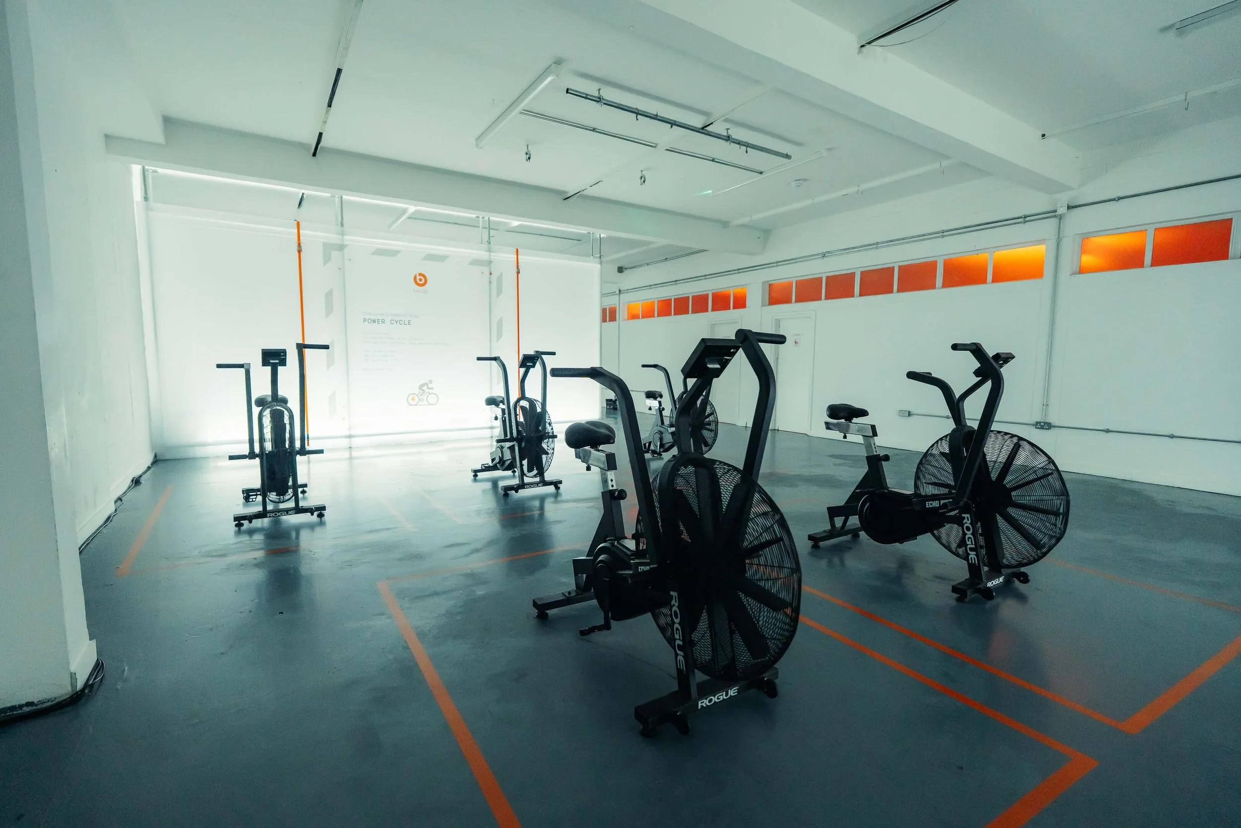 Empty indoor gym with stationary exercise bikes, spaced apart, with orange line markings on the floor and orange accents on windows and wall.