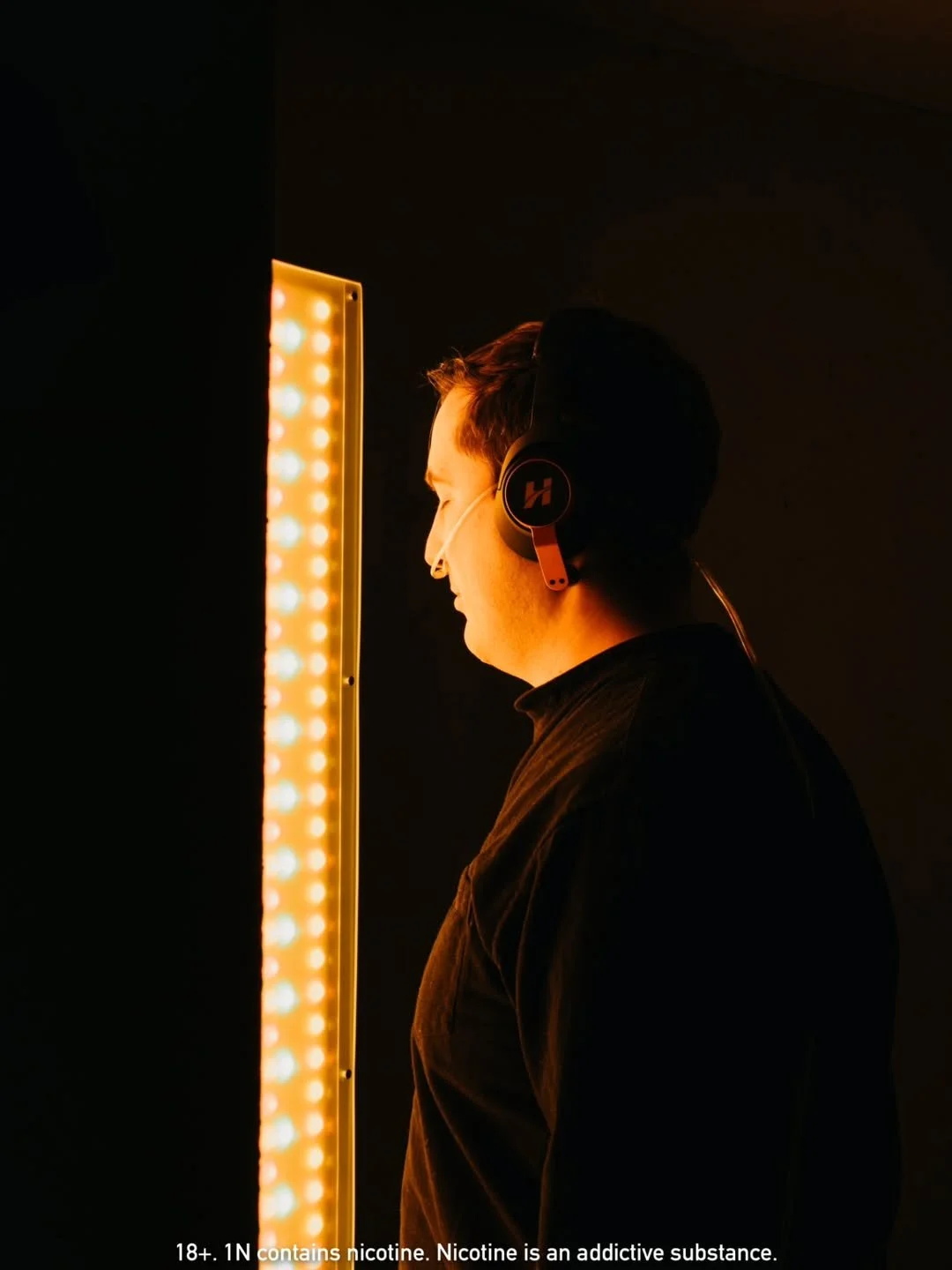 A man wearing headphones and a face mask looking at a vertical panel with orange and white lights in a dark room.