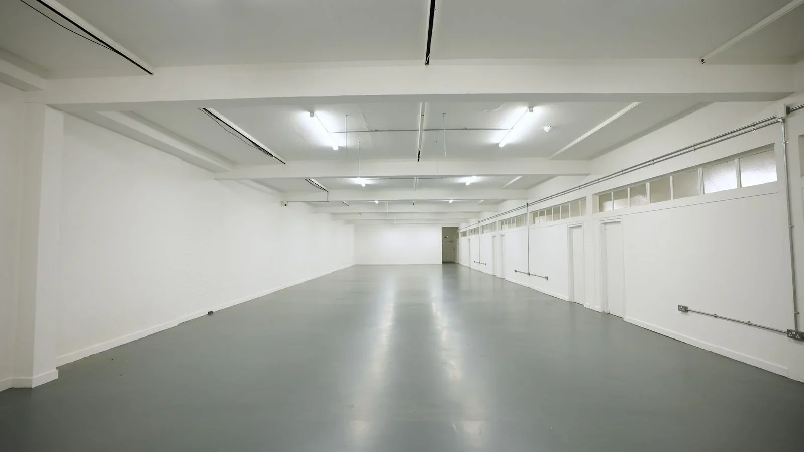 Empty white room with gray flooring, fluorescent lighting, and a series of windows and doors along the walls.