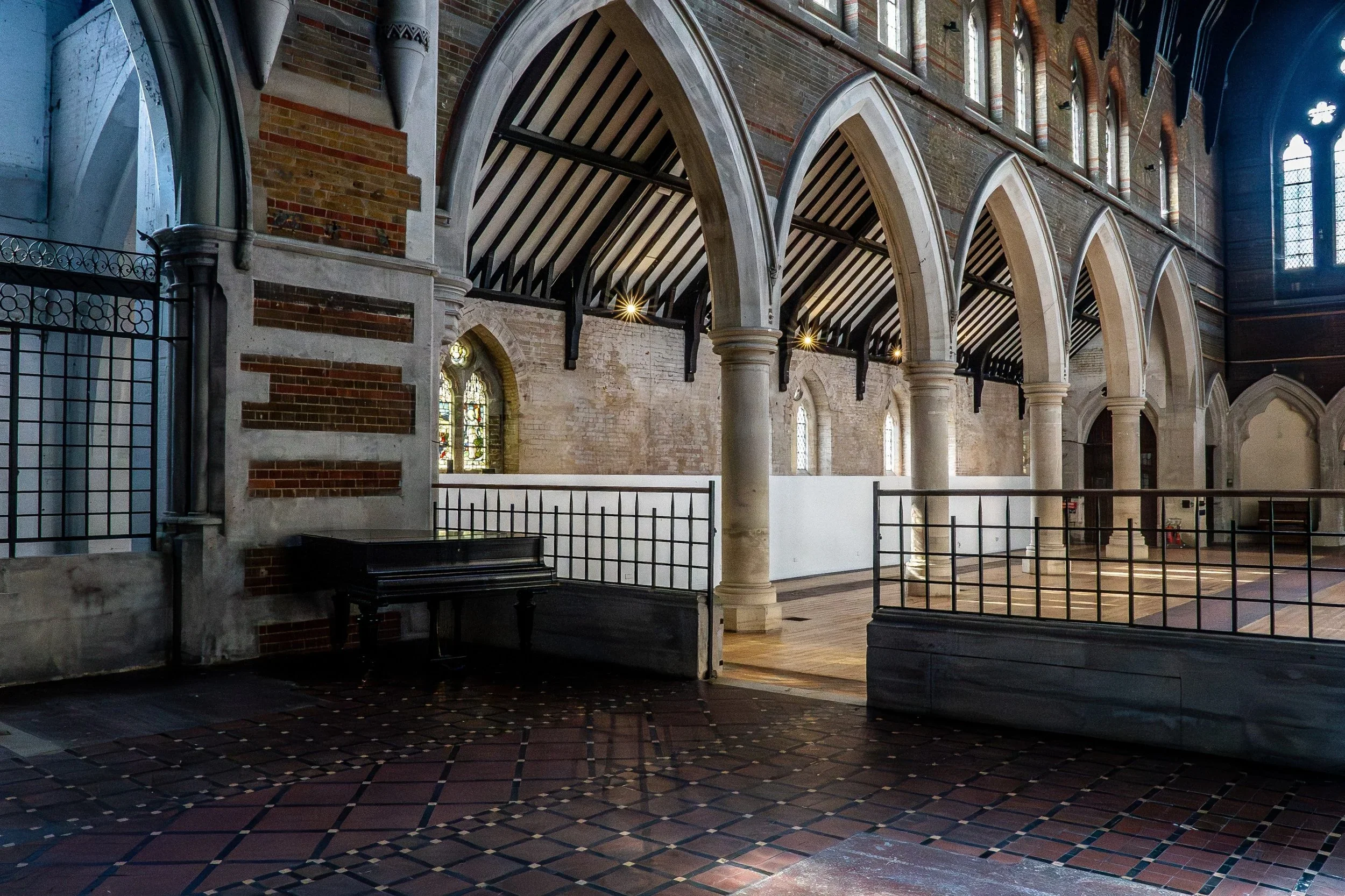 Interior of a church with tall arched windows, stone and brick walls, and a high vaulted ceiling with wooden beams. A black piano is in the corner, and there's a railing separating a section of the floor. The floor is patterned with reddish-brown tiles, and sunlight filters through the stained glass windows.