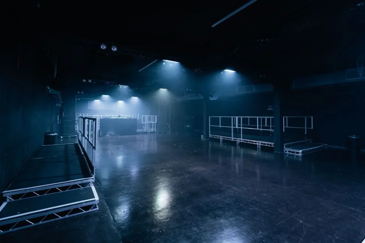 Empty nightclub or event space with dark walls and floors, illuminated by blue overhead lights, with metal stages and railings for performers or DJ setup.