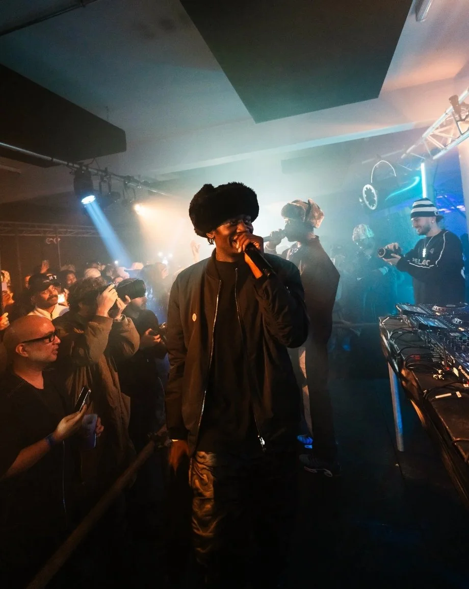 A performer wearing a black fur hat and black jacket singing into a microphone on stage in a club with an audience watching, some taking photos and videos, with DJ equipment and colorful lighting.