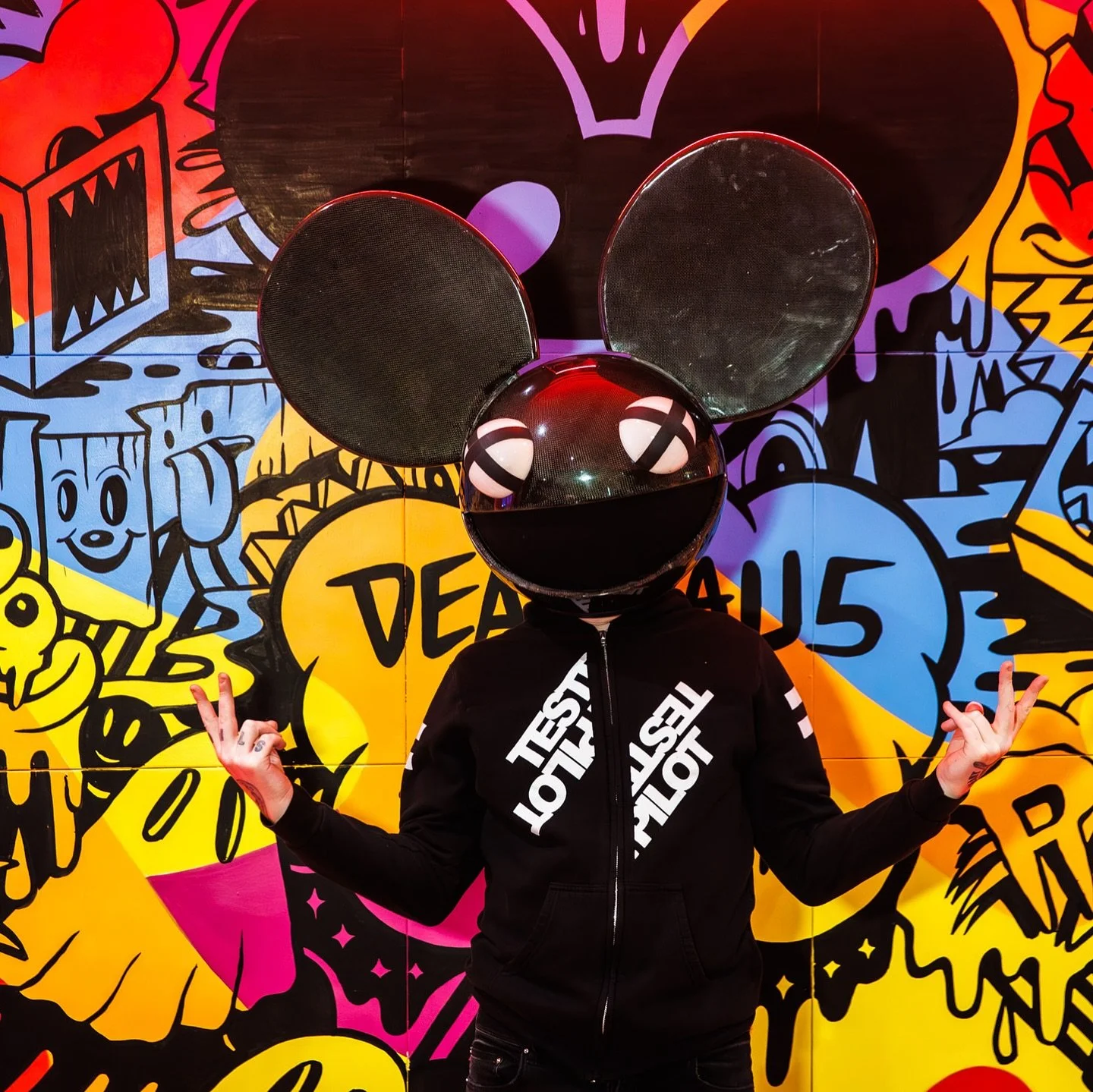 Person wearing a black hoodie with white text and a large helmet with mouse ears and a reflective black sphere for a face, standing in front of a colorful graffiti wall, making hand gestures.