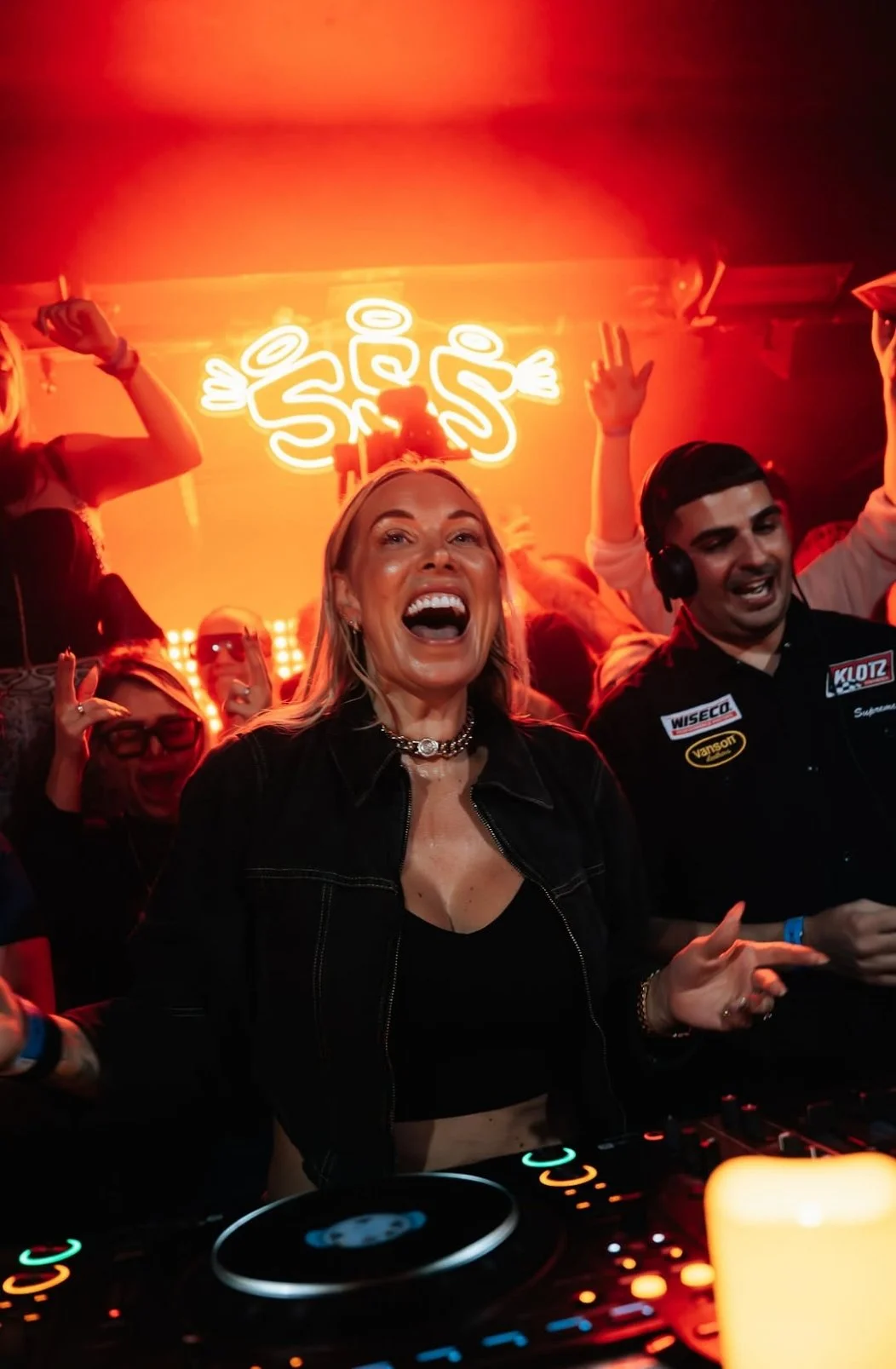 A woman smiling and dancing at a nightclub or party, with a DJ in front of her and people dancing in the background, illuminated by orange neon lighting.