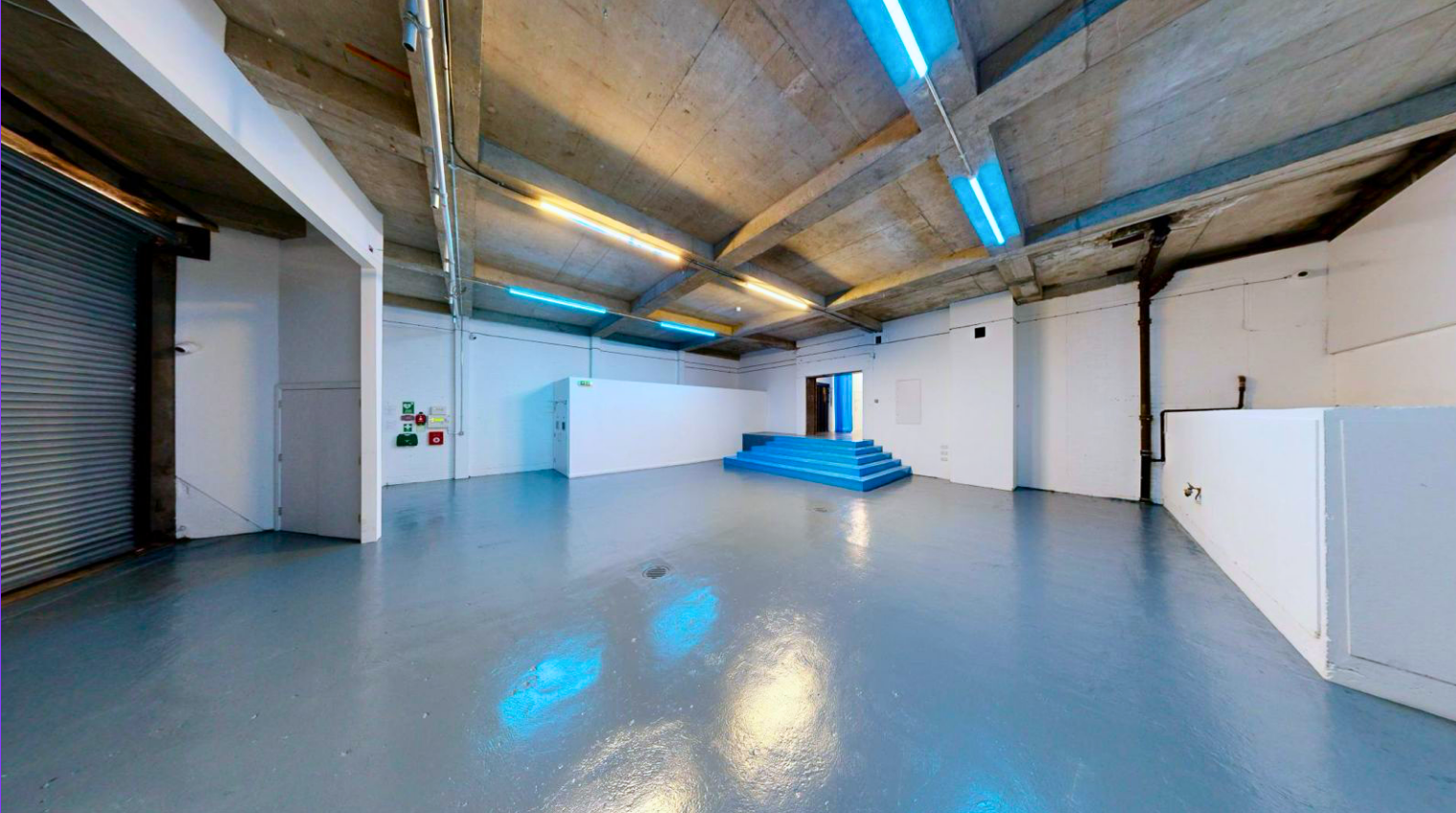 Empty commercial or industrial space with polished concrete floors, white walls, a concrete ceiling with exposed pipes and some blue and yellow lighting fixtures, blue stairs leading up to a doorway, and some safety and fire equipment on the left wall.